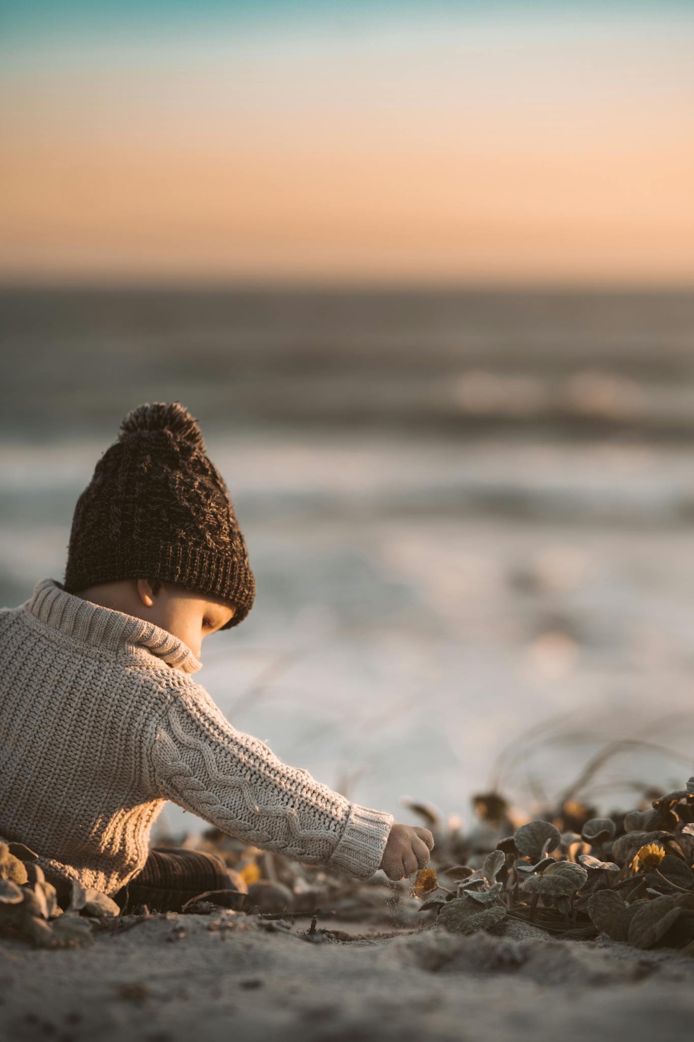 A cute toddler in a knitted sweater playing on a sandy beach at sunset.