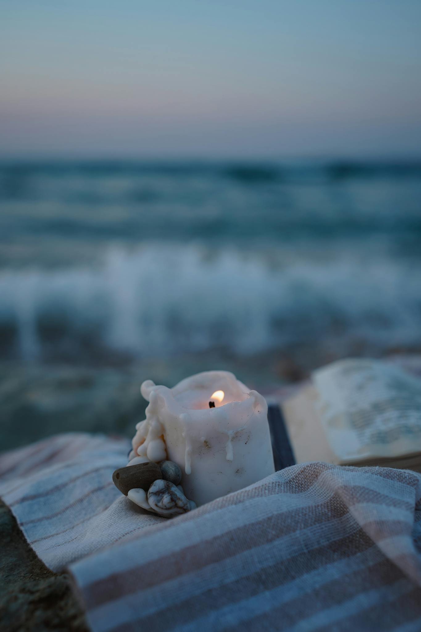 A lit candle on a beach towel by the ocean during twilight, reflecting peace.