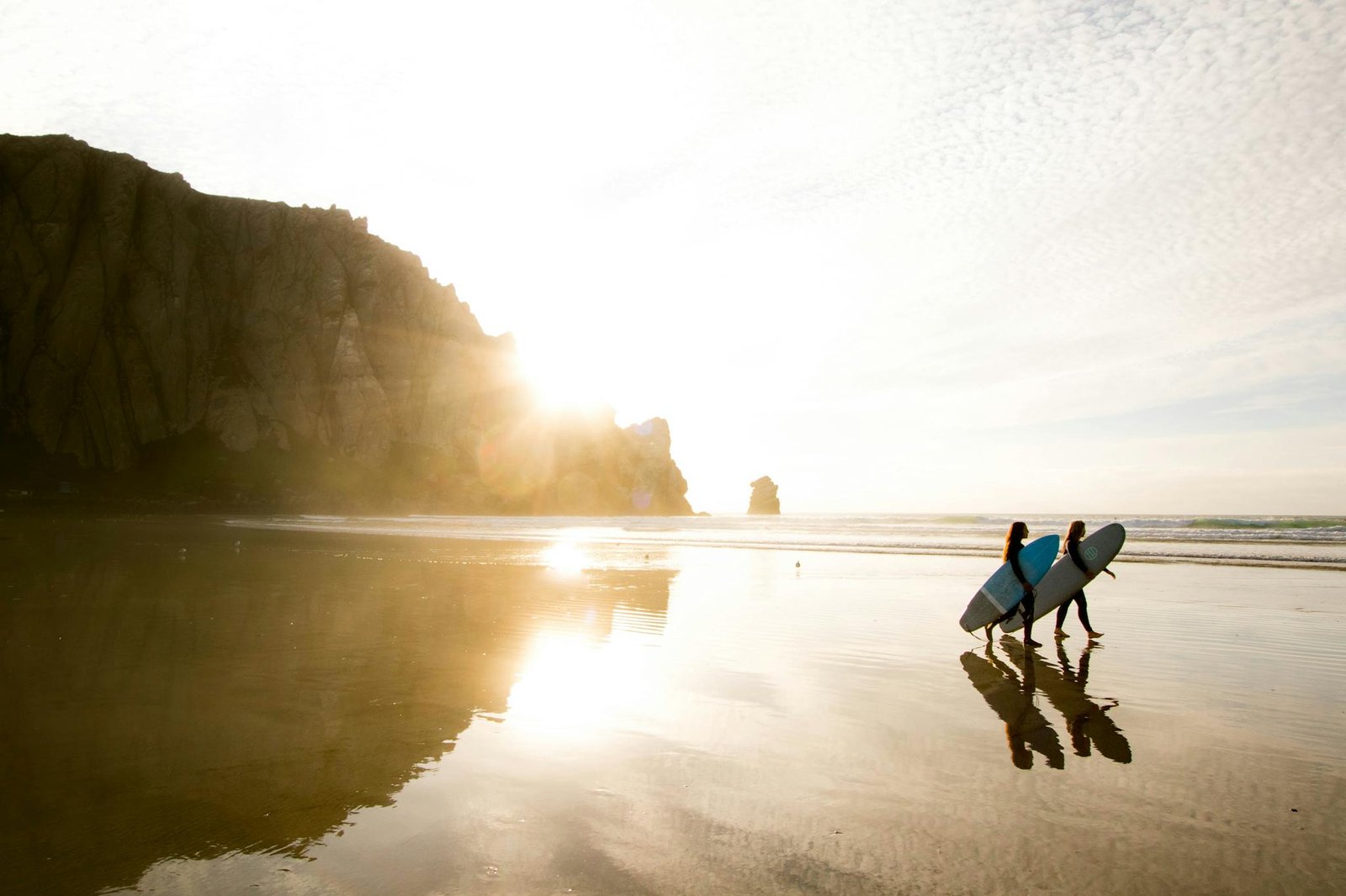 Surfers carrying boards on California beach at sunset with cliffs and ocean reflections.